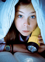 A child in bed under sheets with flashlight reading book